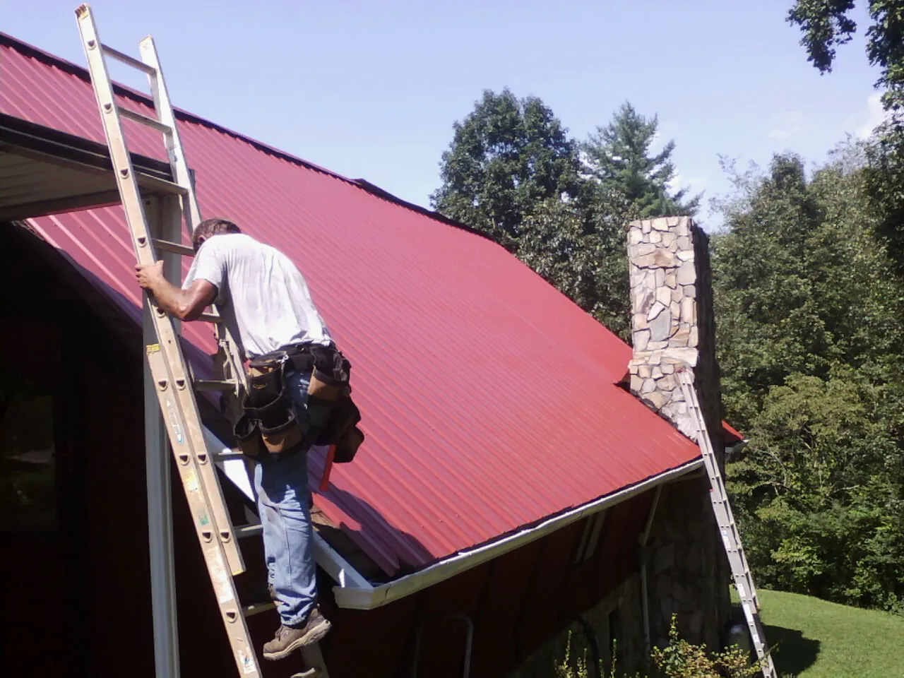 Roofer on a ladder beside a red metal roof and stone chimney — Soffit & Fascia Repair by Roofing Brothers in Morton Grove